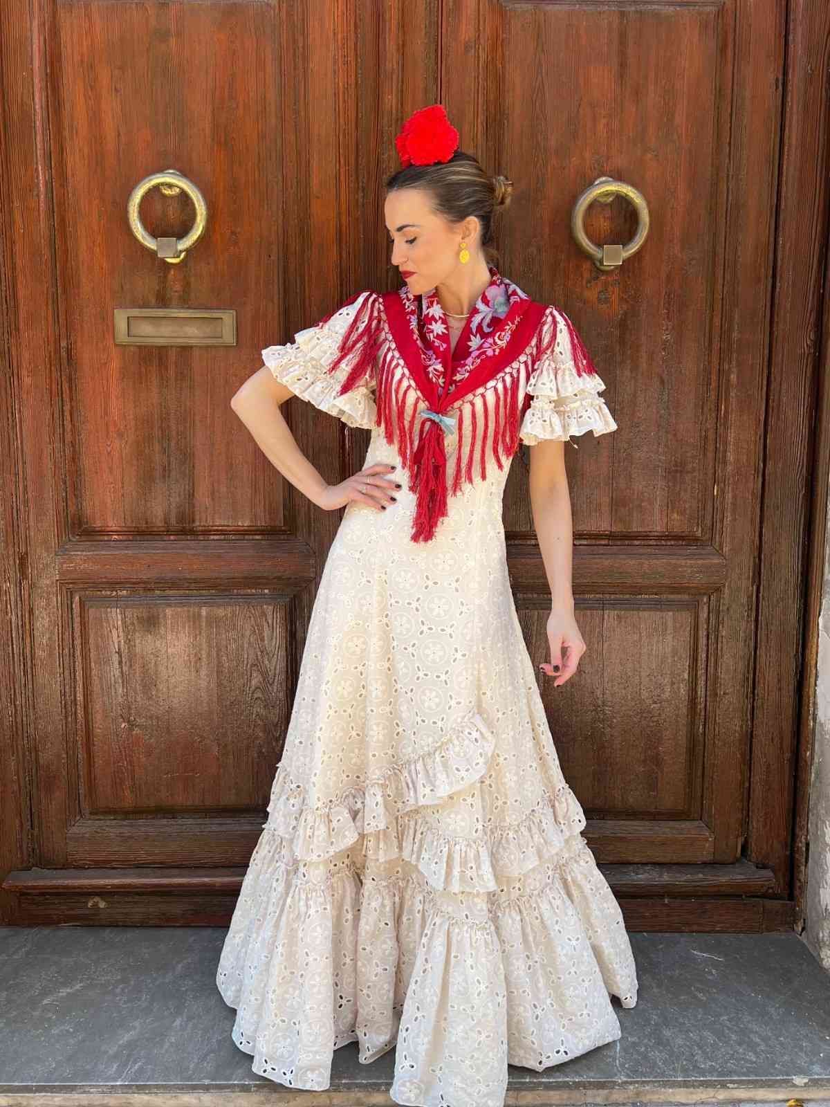 Una persona con un vestido color crema con volantes, un chal rojo y un tocado posa frente a una puerta de madera.
