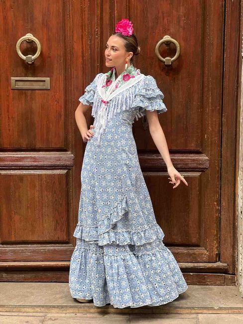 Mujer con un vestido azul con volantes y un tocado de flores rosas, de pie junto a una puerta de madera.