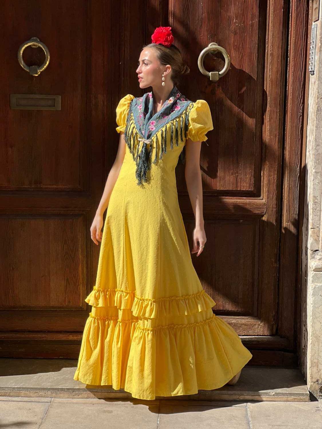 Una persona con un vestido amarillo brillante con volantes está de pie junto a una puerta de madera con anillos de latón y un accesorio rojo para el cabello.