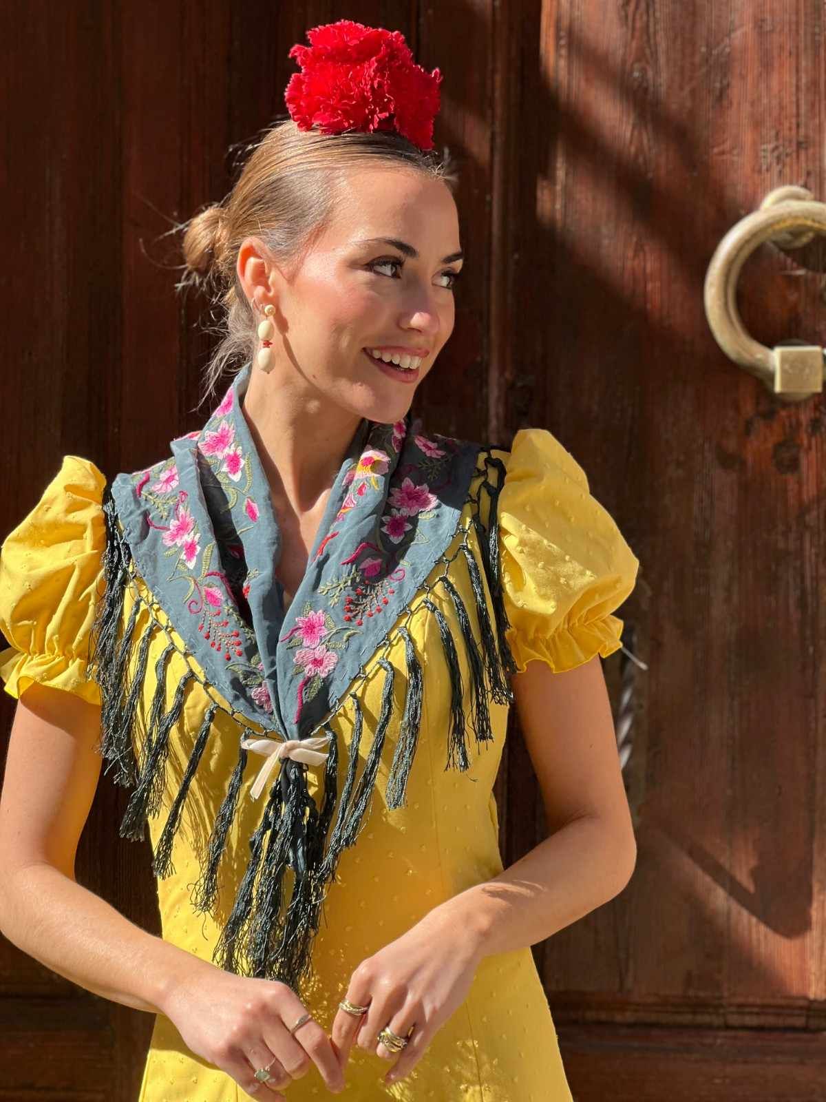 Mujer con vestido amarillo, pañuelo gris y tocado de flores rojas, de pie junto a una puerta de madera.