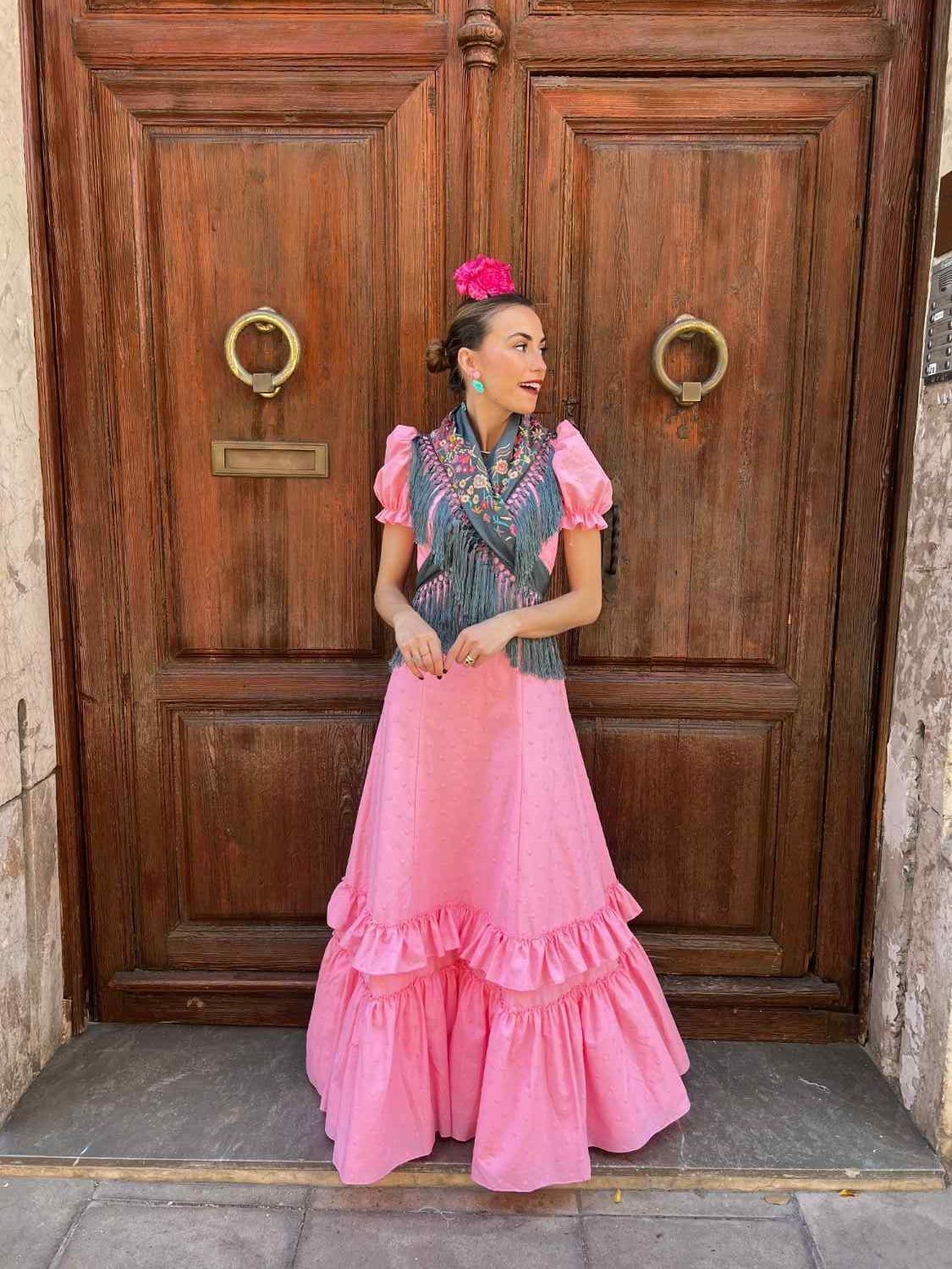 Mujer con vestido rosa de pie frente a una gran puerta de madera con anillos de latón.