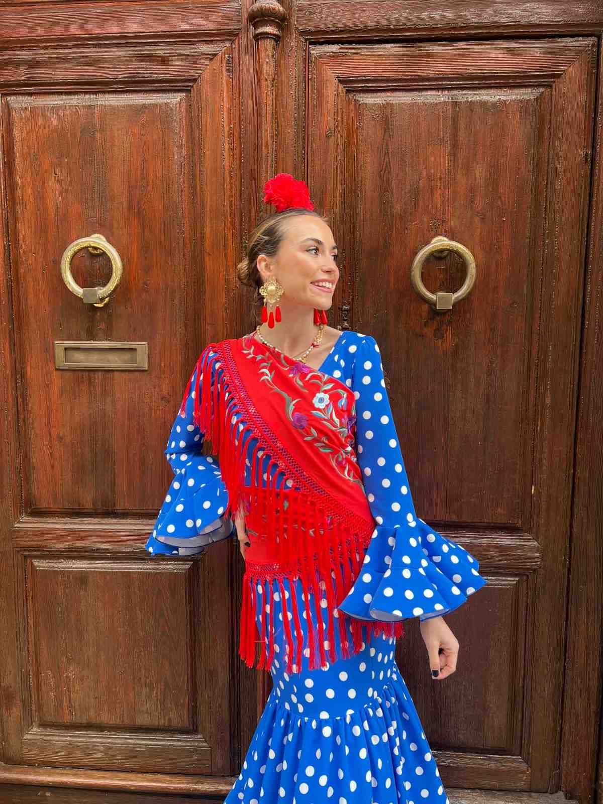 Mujer con un vestido azul de lunares con flecos rojos posando frente a unas puertas de madera tallada.