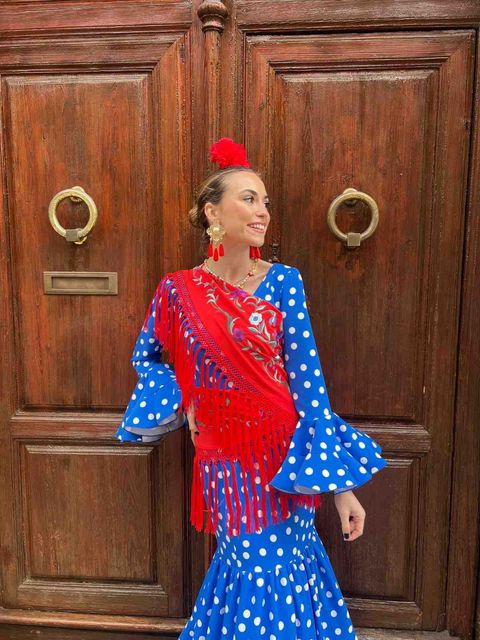 Mujer con un vestido azul de lunares con flecos rojos posando frente a una puerta de madera.