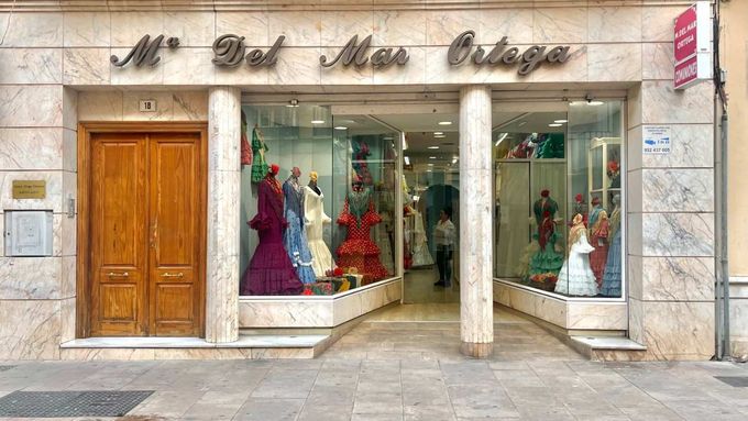 Fachada de una tienda de vestidos de novia con maniquíes con vestidos coloridos y una puerta de madera a la izquierda.