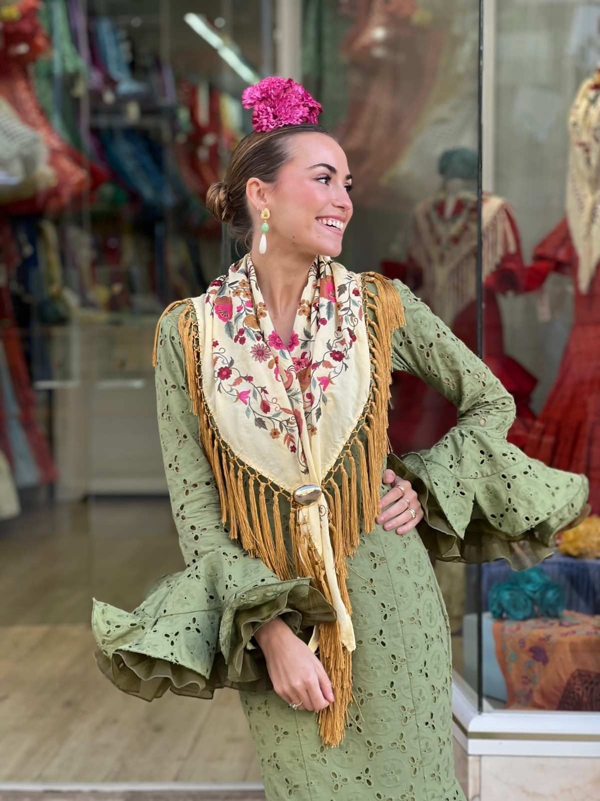 Una persona vestida con un atuendo verde, una bufanda floral y un tocado rosa posa en el escaparate de una tienda.