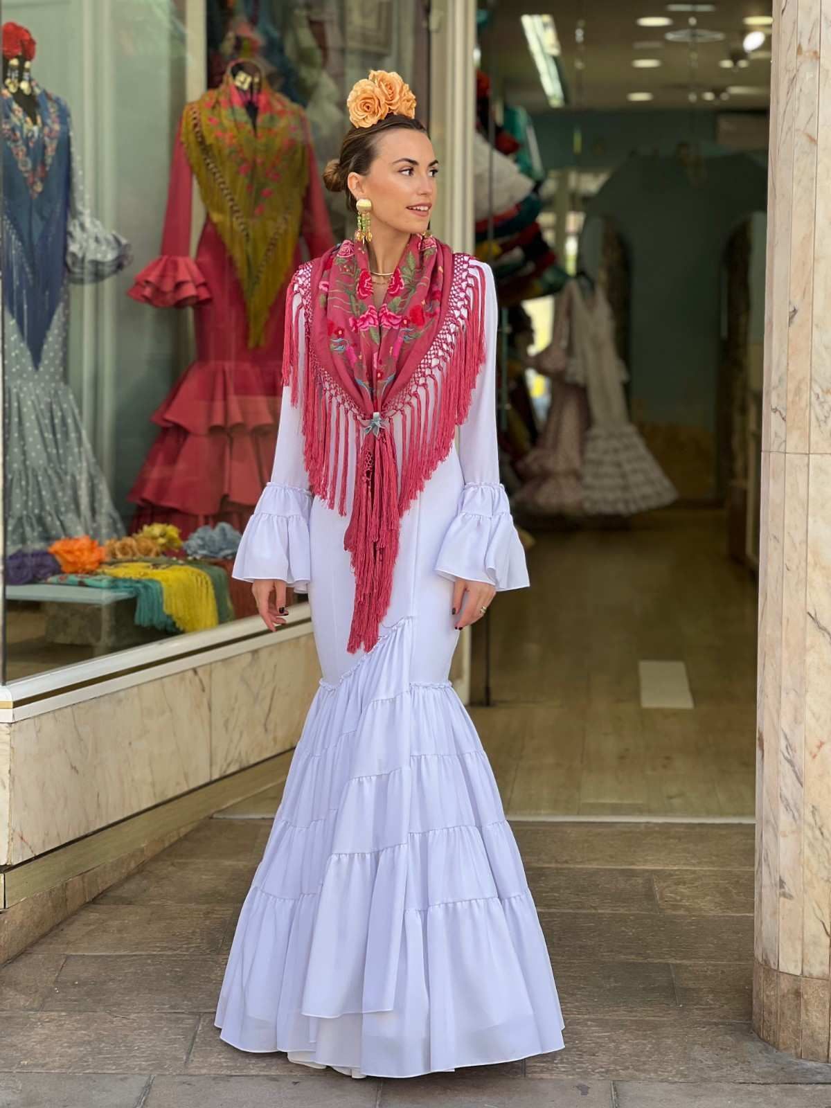 Mujer con vestido blanco y chal rojo estampado de pie en la entrada de una boutique.