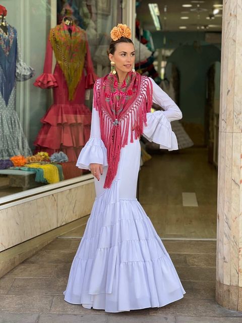 Mujer con vestido blanco con volantes y chal rosa posando en una boutique de ropa.