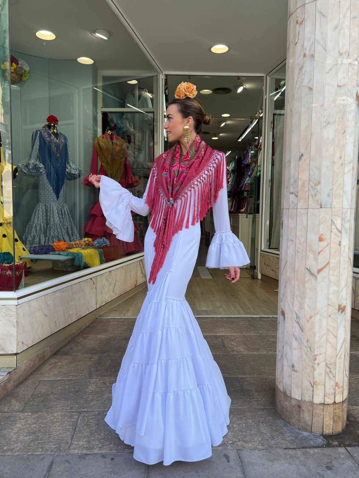 Una persona vestida con un vestido blanco y un chal rosa está de pie frente al escaparate de una tienda de ropa.