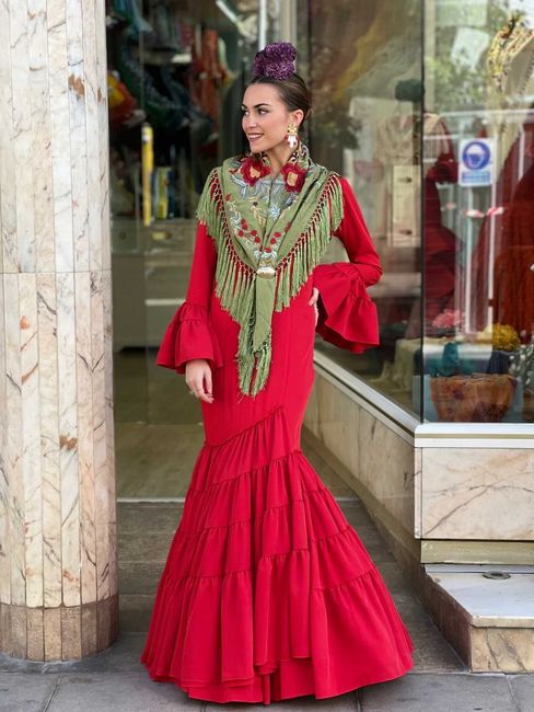 Mujer con un vestido rojo brillante con volantes y elaborados adornos verdes de pie frente a una tienda.