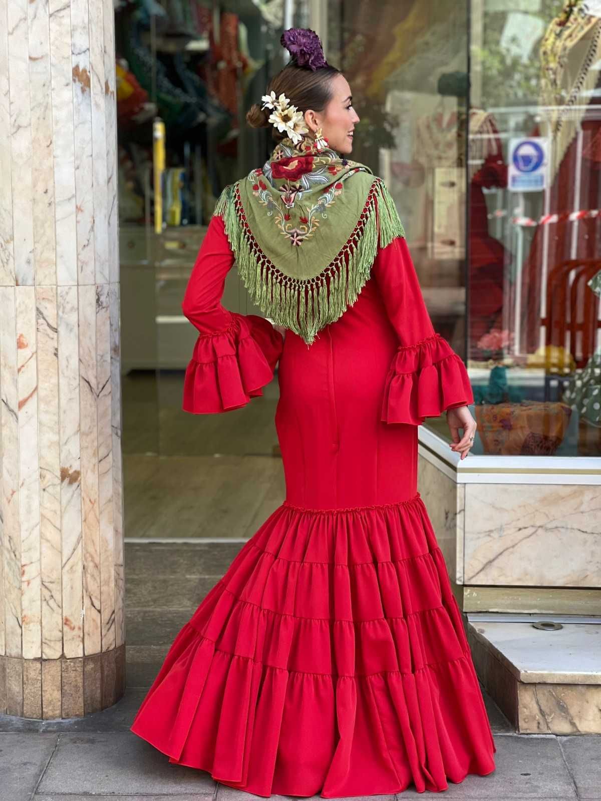Mujer con un vestido rojo con volantes y un chal verde, de pie cerca de la entrada de un edificio.