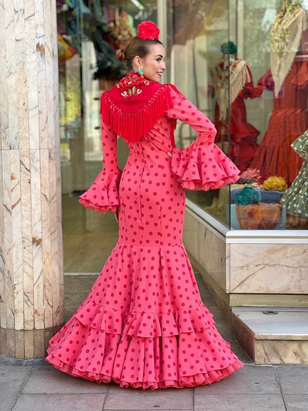 Mujer con un vestido rosa brillante con volantes posando junto al escaparate de una tienda.