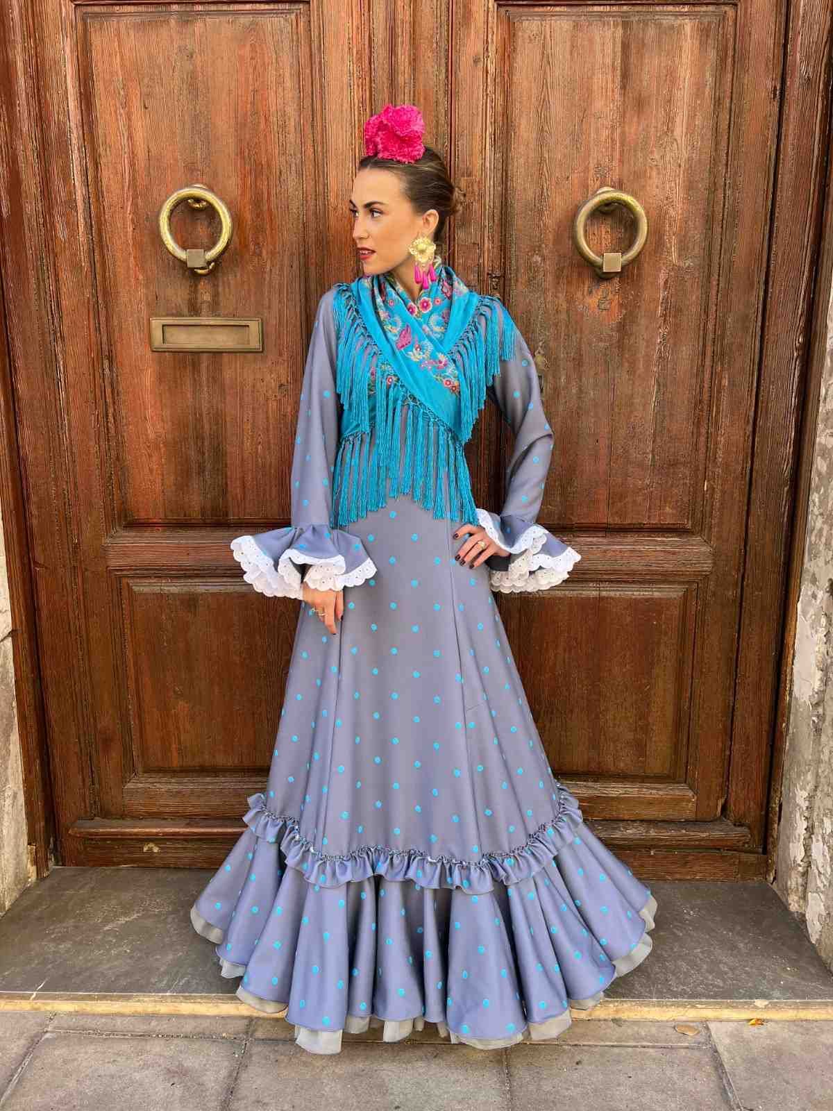 Mujer con un traje tradicional azul y gris de pie frente a unas puertas dobles de madera.