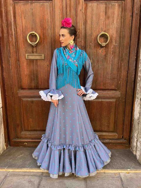 Persona con vestido verde azulado y gris posando frente a una puerta de madera con anillos de latón.