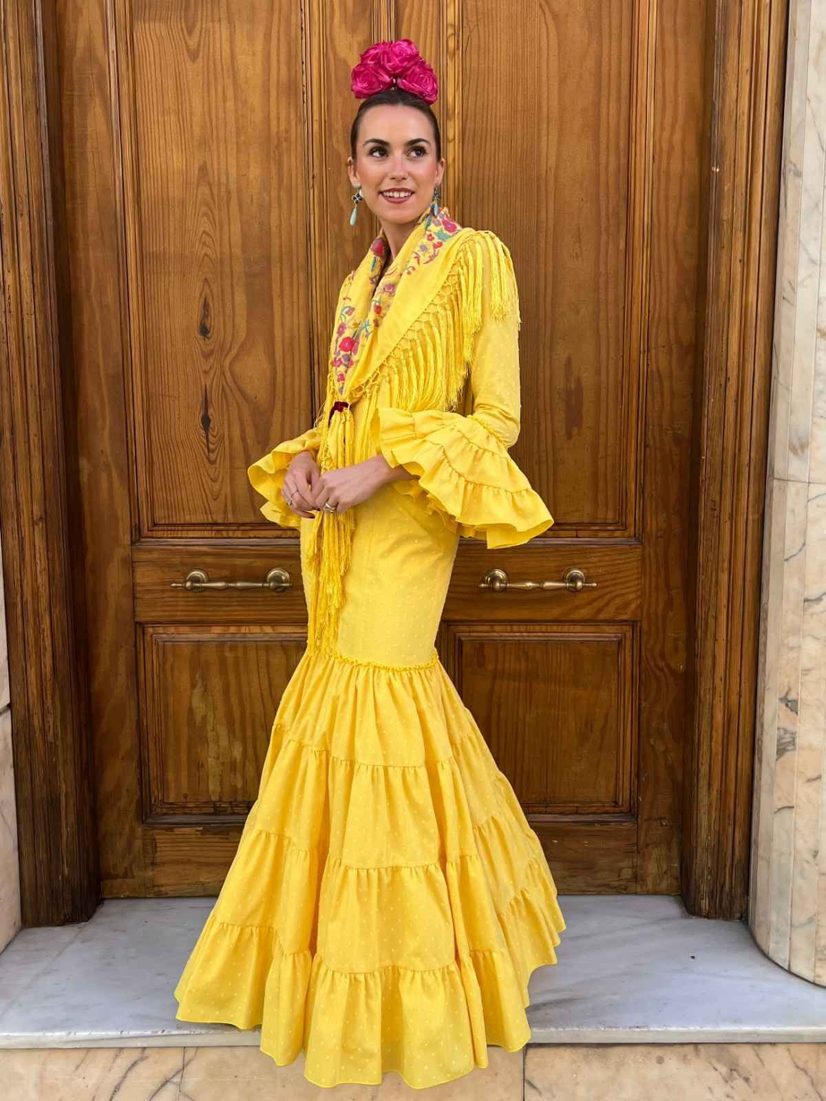 Mujer con un vestido amarillo brillante con volantes posando frente a una puerta de madera, luciendo un tocado floral rosa.