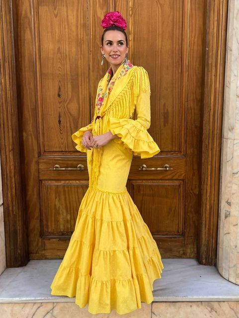 Mujer con un vestido amarillo brillante con volantes posando frente a una puerta de madera, luciendo un tocado floral rosa.