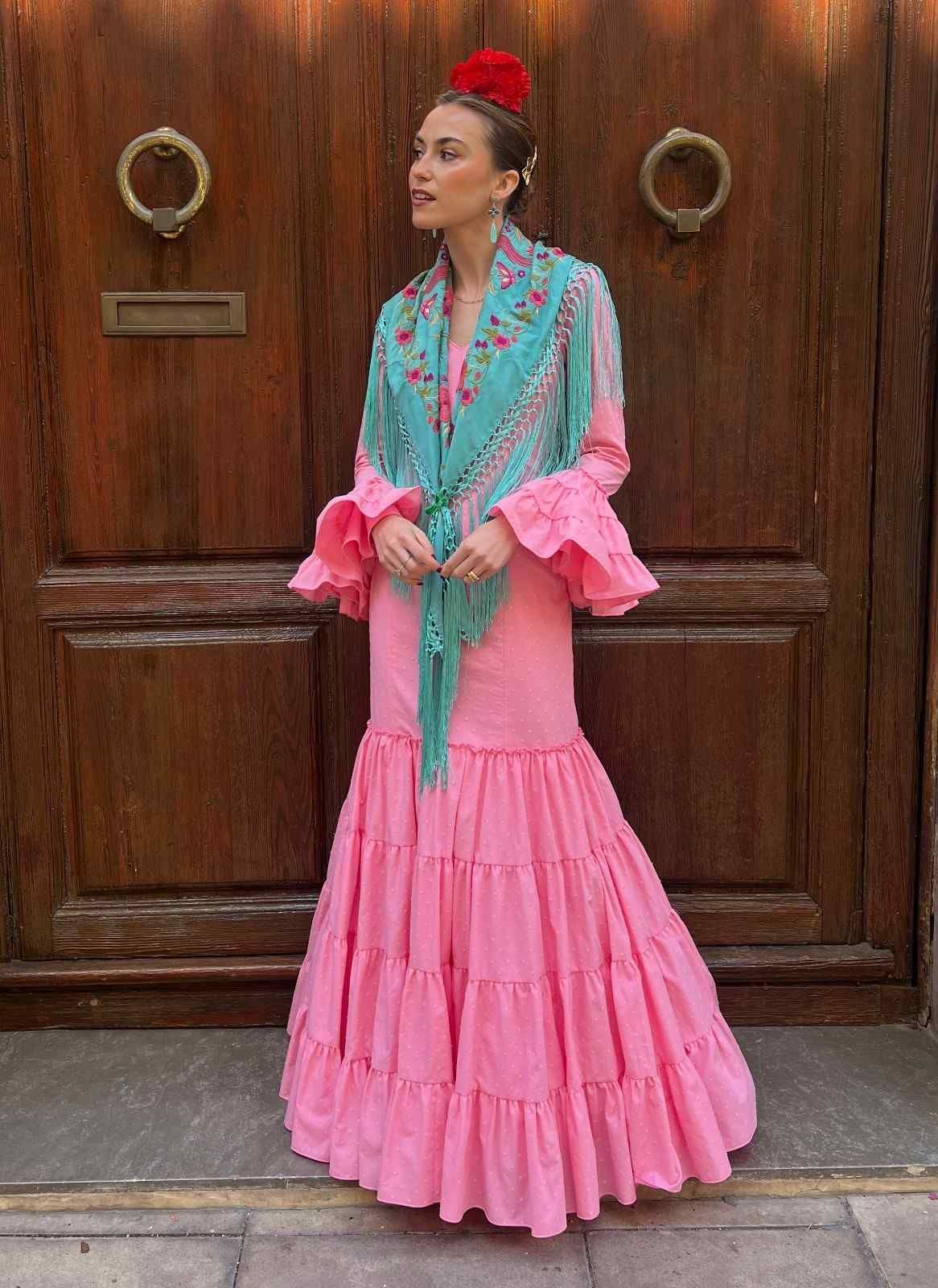 Mujer con un vestido rosa de volantes, bufanda turquesa y mangas rosas con volantes, de pie frente a una puerta de madera.