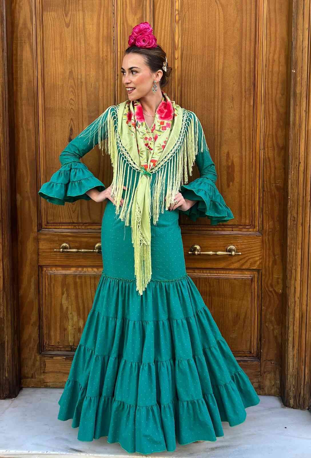 Mujer con un vestido turquesa con volantes y detalles florales, posando frente a unas puertas de madera.