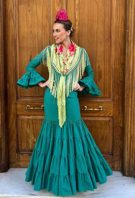 Mujer con un vestido turquesa con volantes y detalles florales, posando frente a unas puertas de madera.