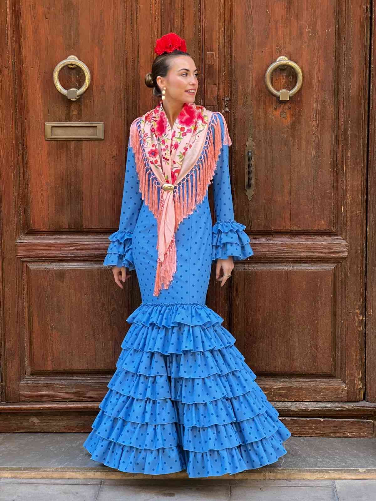 Mujer con un vestido azul con volantes, de pie junto a una puerta de madera, mirando hacia un lado.