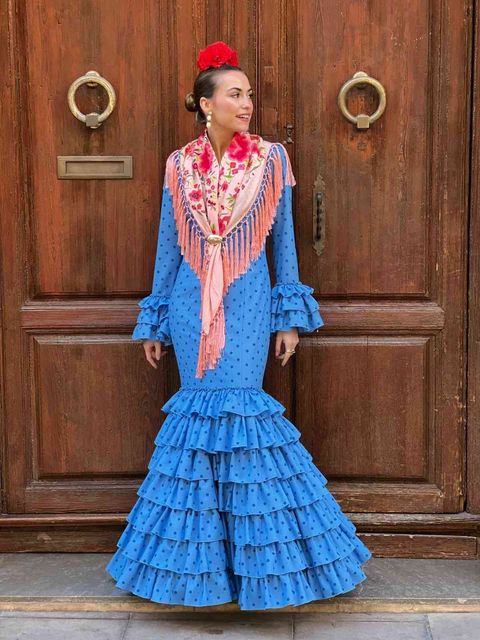 Mujer con un vestido azul con volantes de pie junto a una puerta de madera con tiradores circulares.