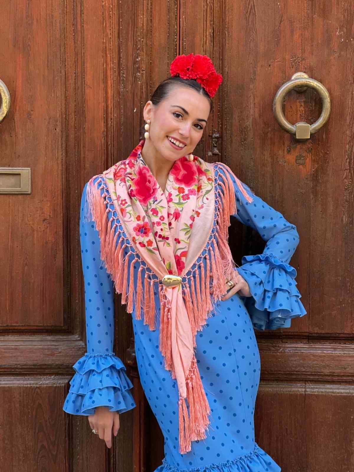 Mujer con un vestido azul de lunares posando junto a una puerta de madera, con un tocado de flores rojas y una bufanda rosa.