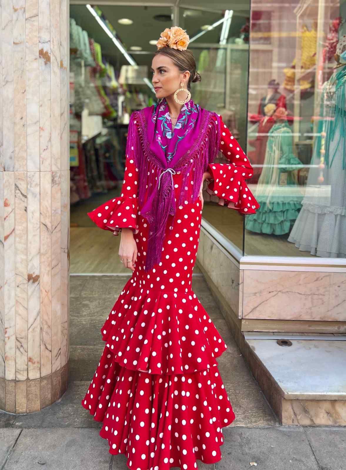 Mujer con un vestido rojo de lunares con volantes, de pie frente al escaparate de una tienda, con una bufanda morada.