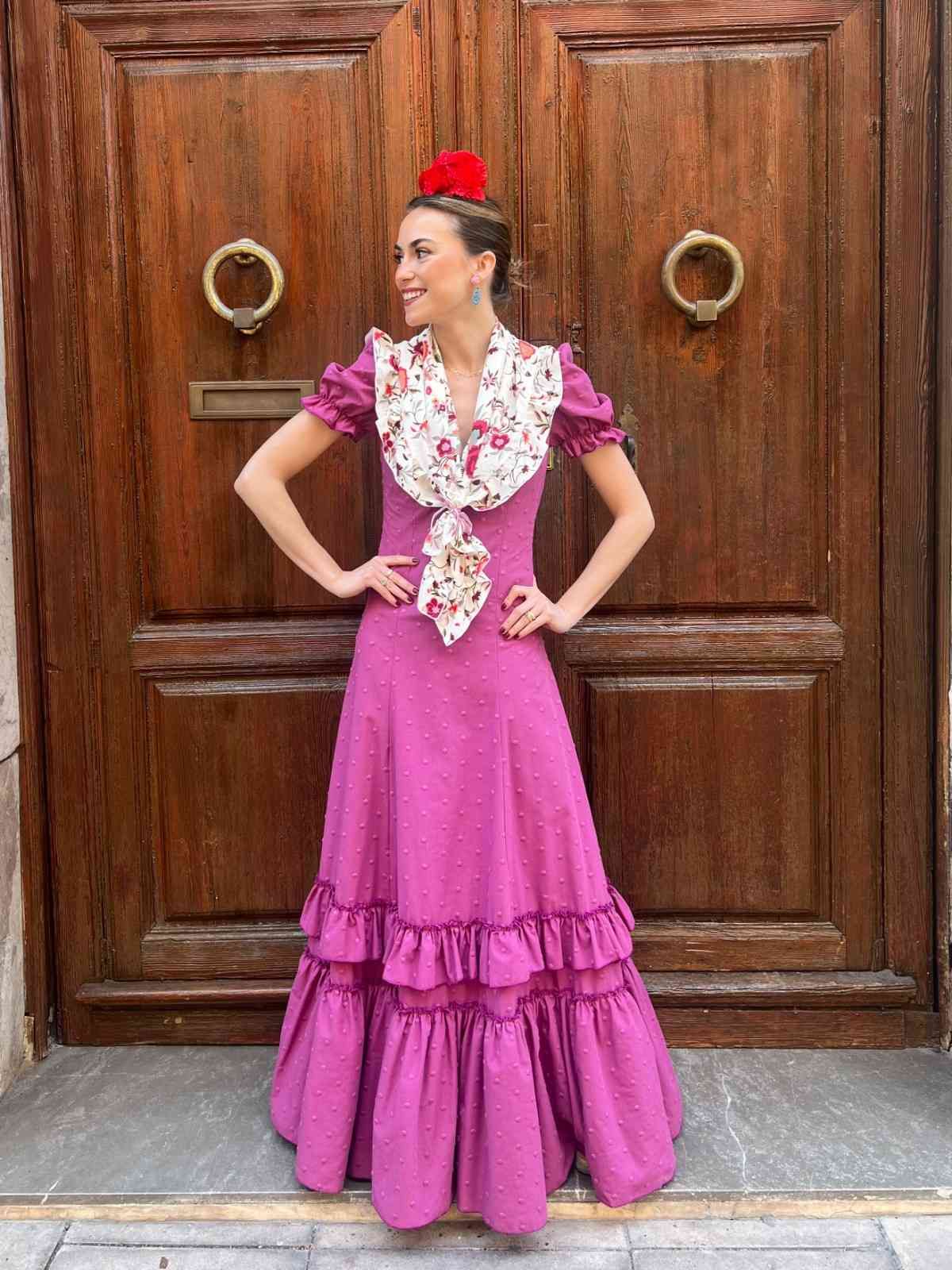 Mujer con un vestido rosa con volantes posando frente a una puerta de madera con anillas de latón.