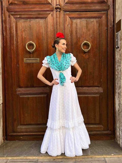 Mujer con vestido blanco, chal turquesa y flor roja, de pie frente a unas grandes puertas de madera.