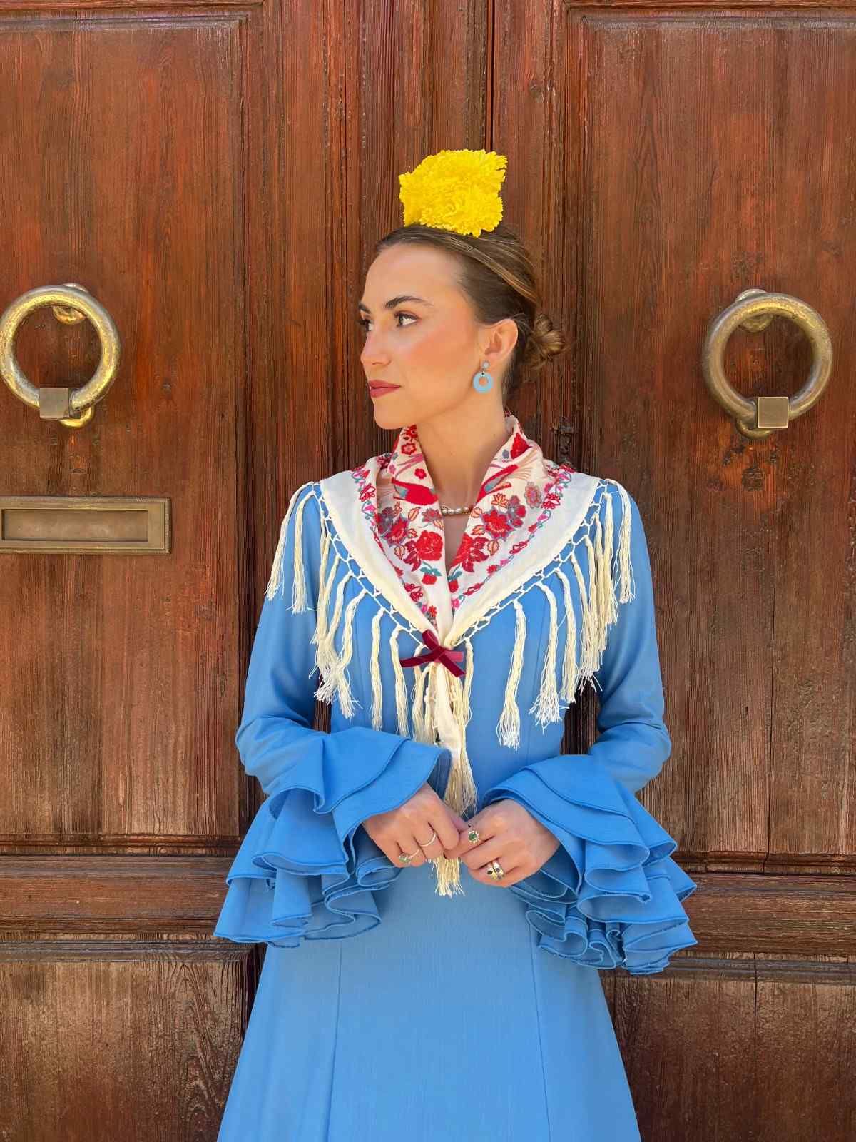 Mujer con un vestido azul bordado de pie frente a una puerta de madera, con una flor amarilla en el cabello.