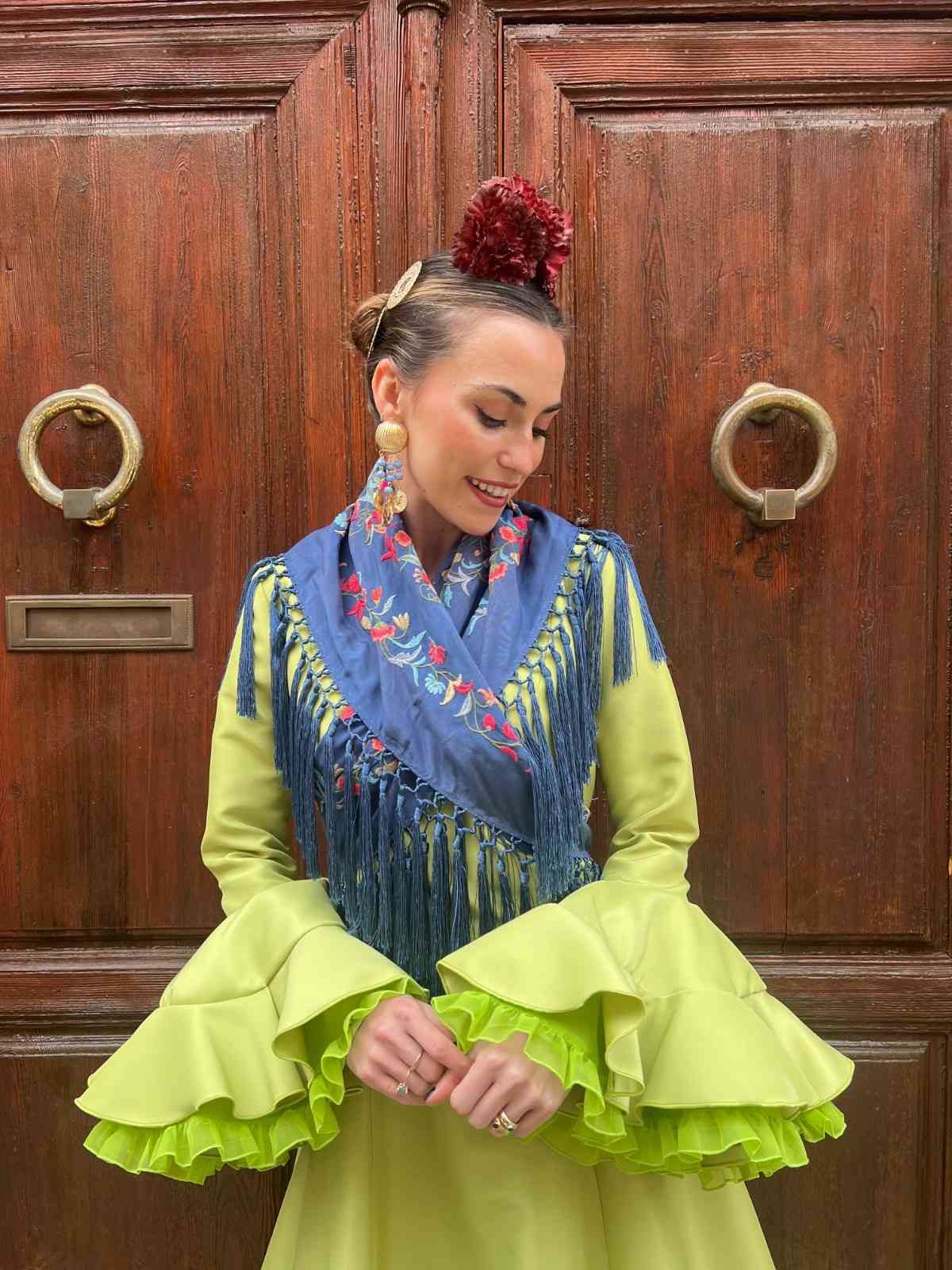 Mujer con un vestido verde lima de pie frente a unas puertas de madera, mirando hacia abajo con las manos entrelazadas.