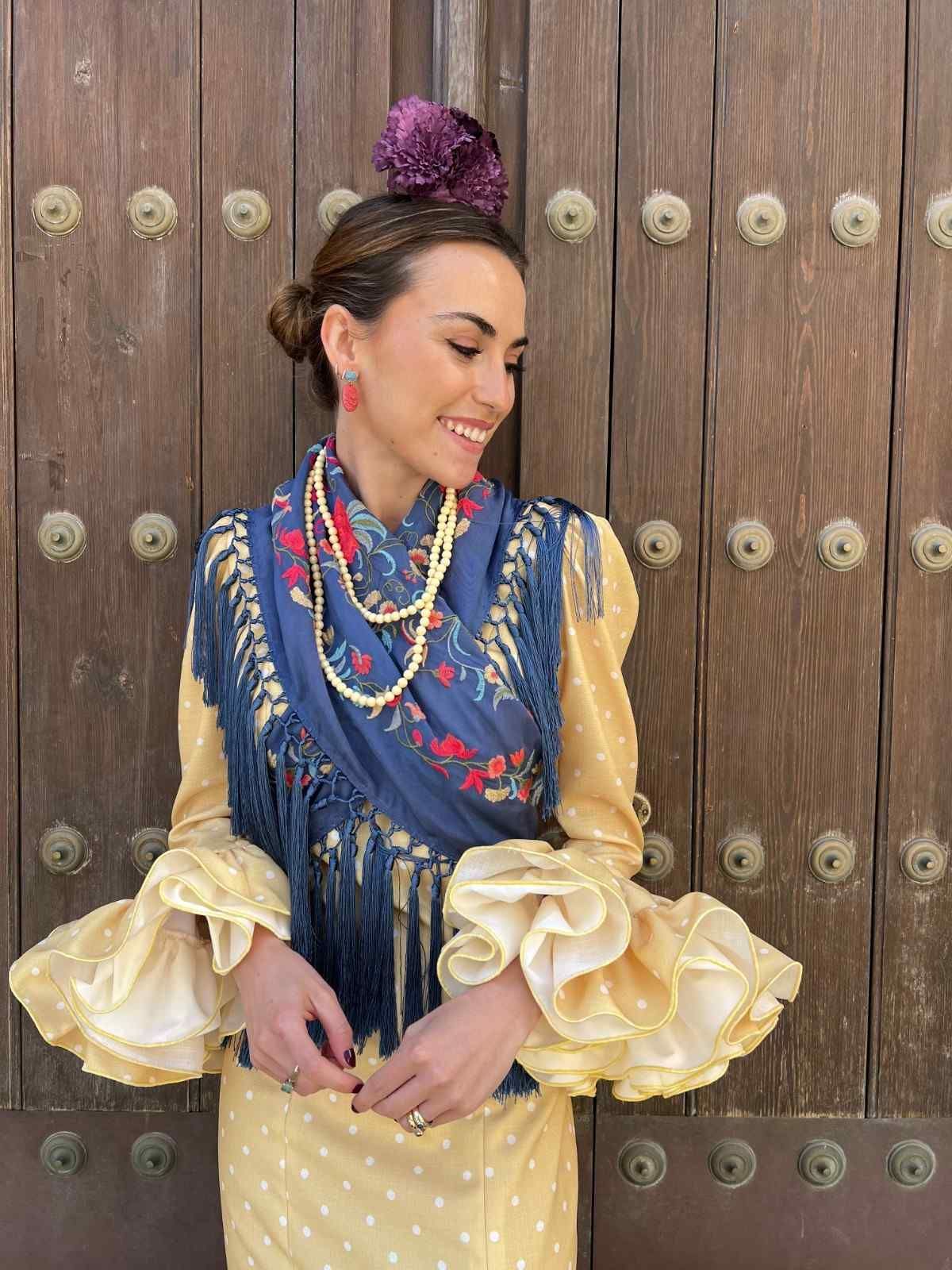Una persona vestida con un traje tradicional azul y dorado se encuentra de pie frente a una puerta de madera tallada, mirando hacia abajo con una sonrisa.