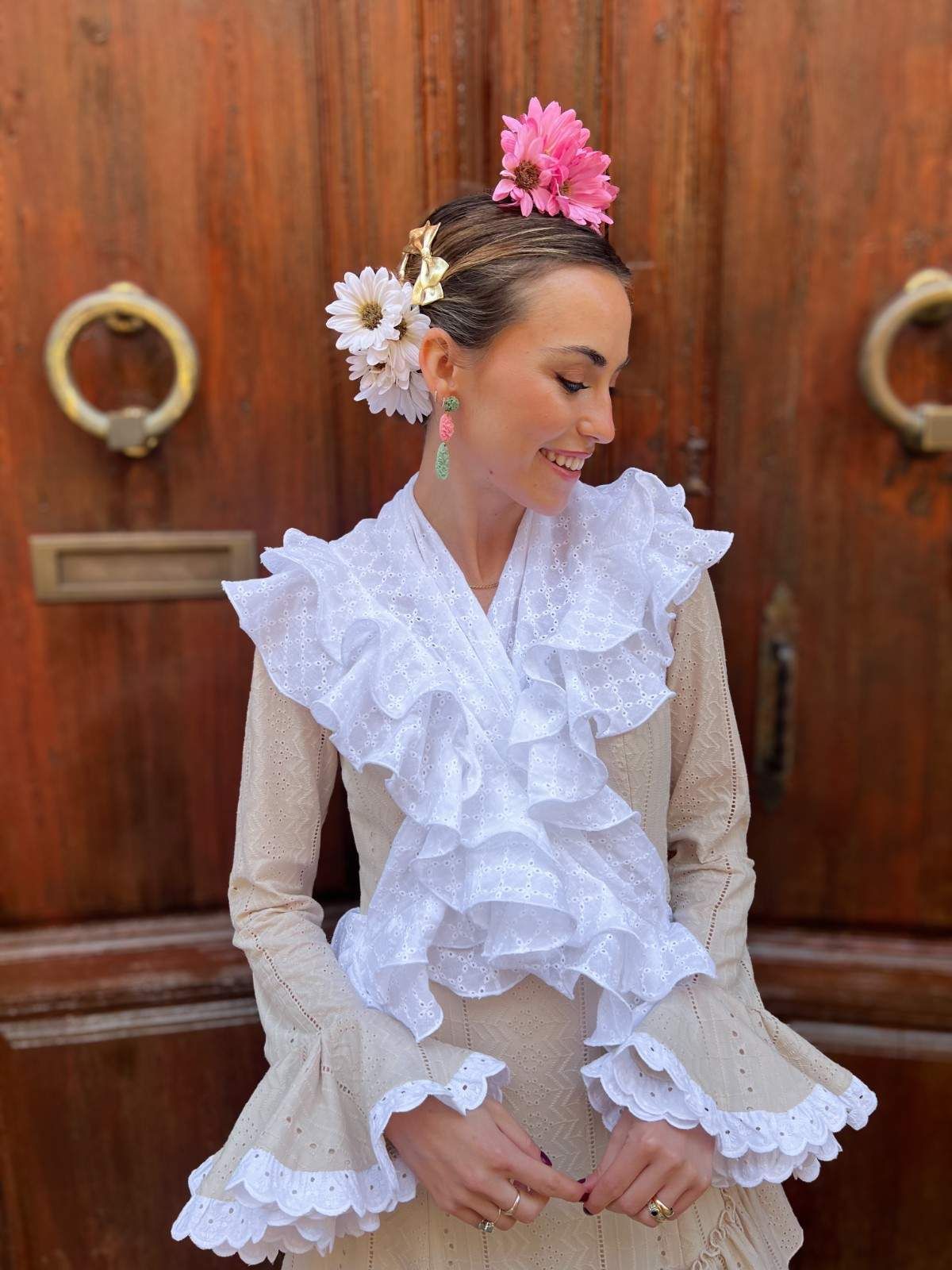 Mujer con un vestido blanco con volantes y flores en el pelo, de pie frente a una puerta de madera con anillas de latón.