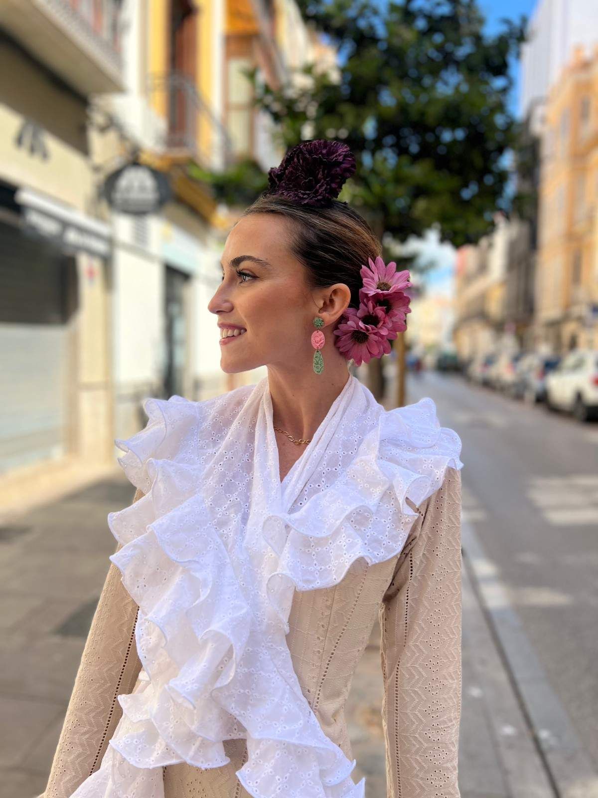 Mujer con blusa blanca con volantes y flores rosas en el pelo, de pie en una calle soleada.