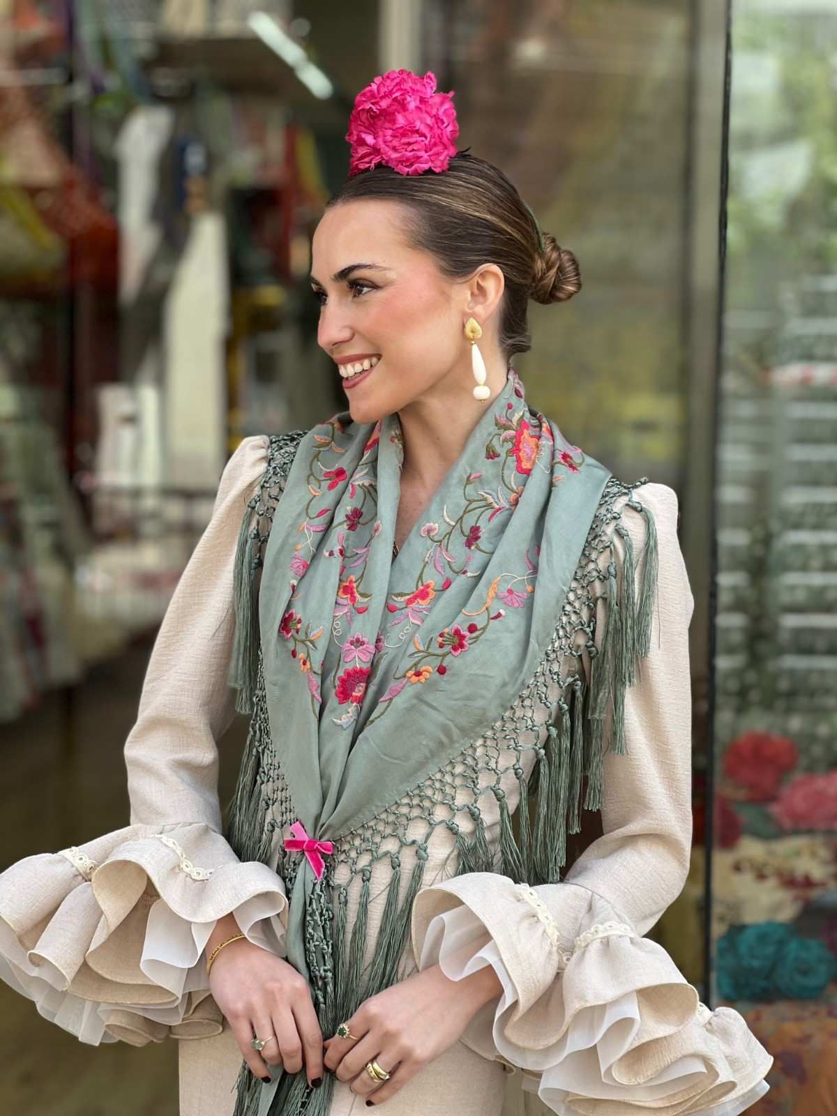 Mujer con un vestido beige con volantes y un chal menta, sonriendo al aire libre con una flor rosa brillante en el cabello.