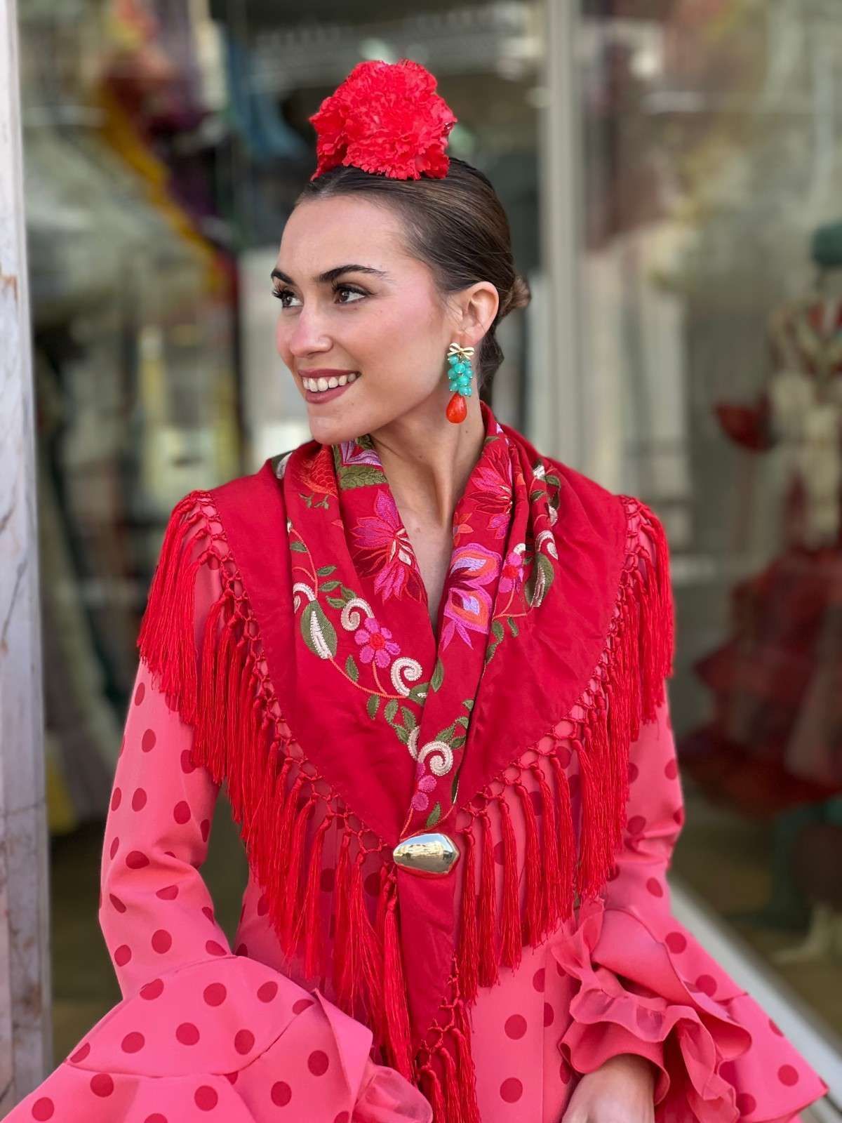 Mujer con un vestido rosa de lunares, un chal rojo con flecos y un accesorio floral para el cabello, sonriendo al aire libre.