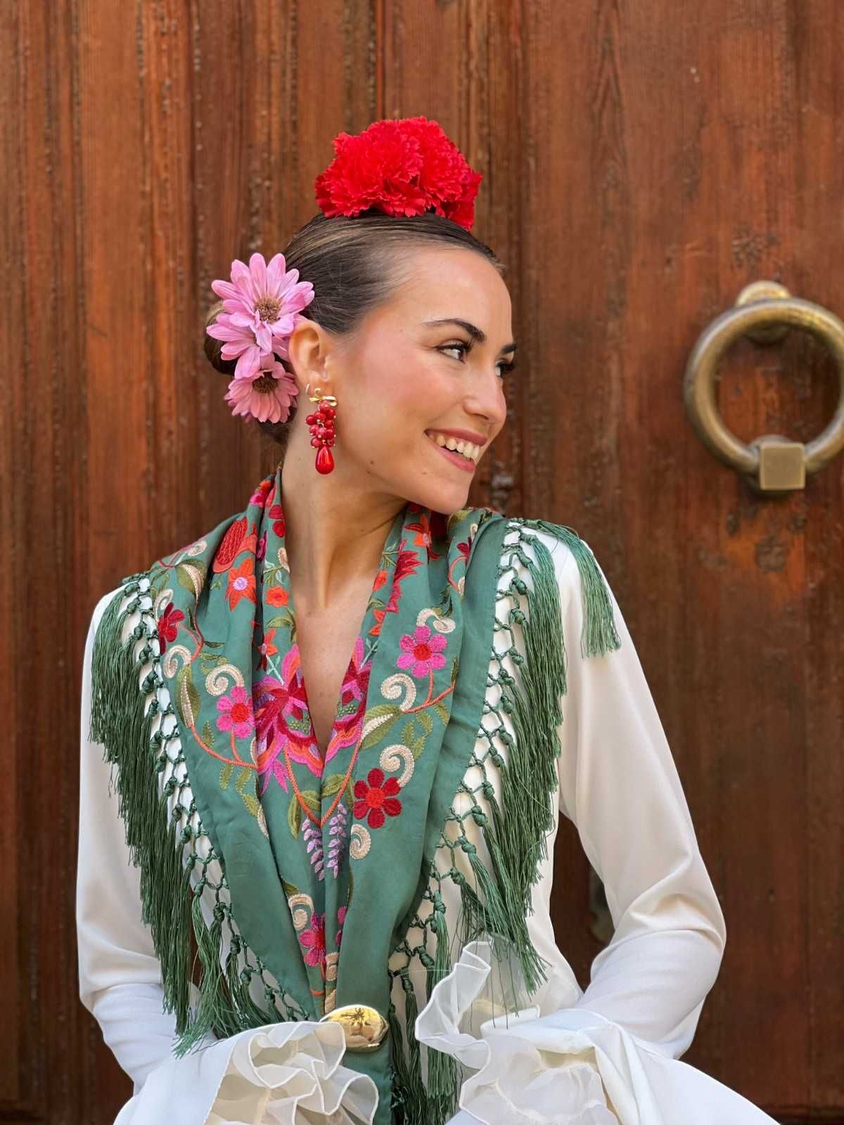 Mujer con chal estampado de flores y vestido blanco, sonriendo de perfil contra una puerta de madera con aldaba de metal.