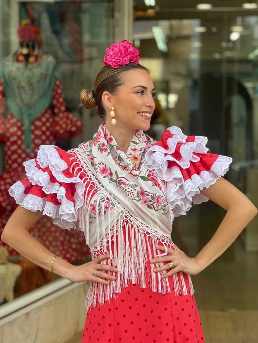 Mujer con un vestido rojo de flamenco, mangas con volantes y tocado floral, posando frente a una tienda.