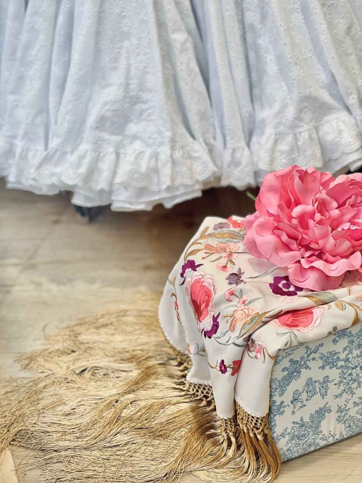 Una flor rosa sobre un taburete acolchado con estampado floral, junto a un vestido blanco y una alfombra beige de pelo largo.