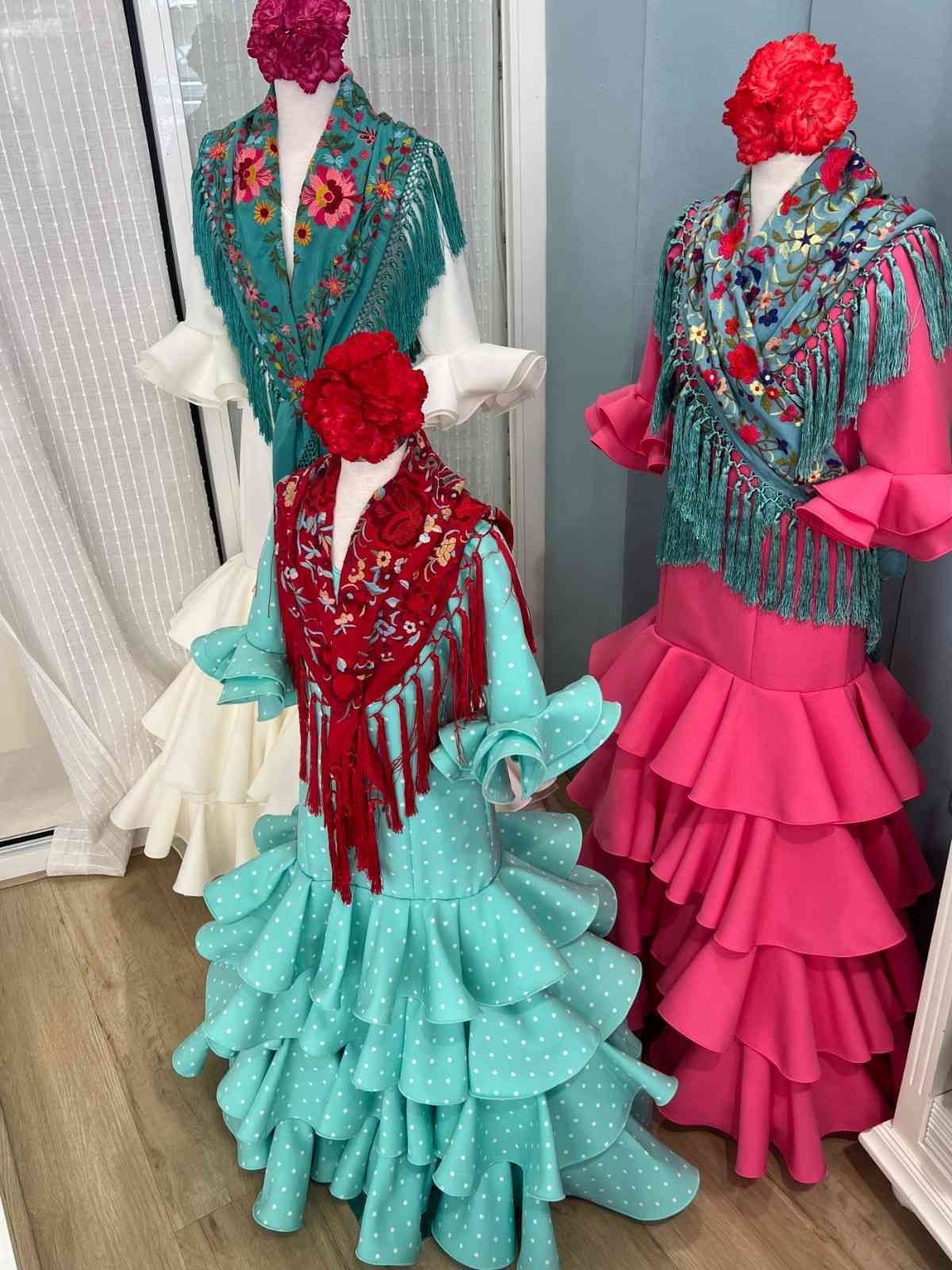 Tres vestidos de estilo flamenco en maniquíes, en color verde azulado, rosa y crema, con faldas con volantes y tocados florales.