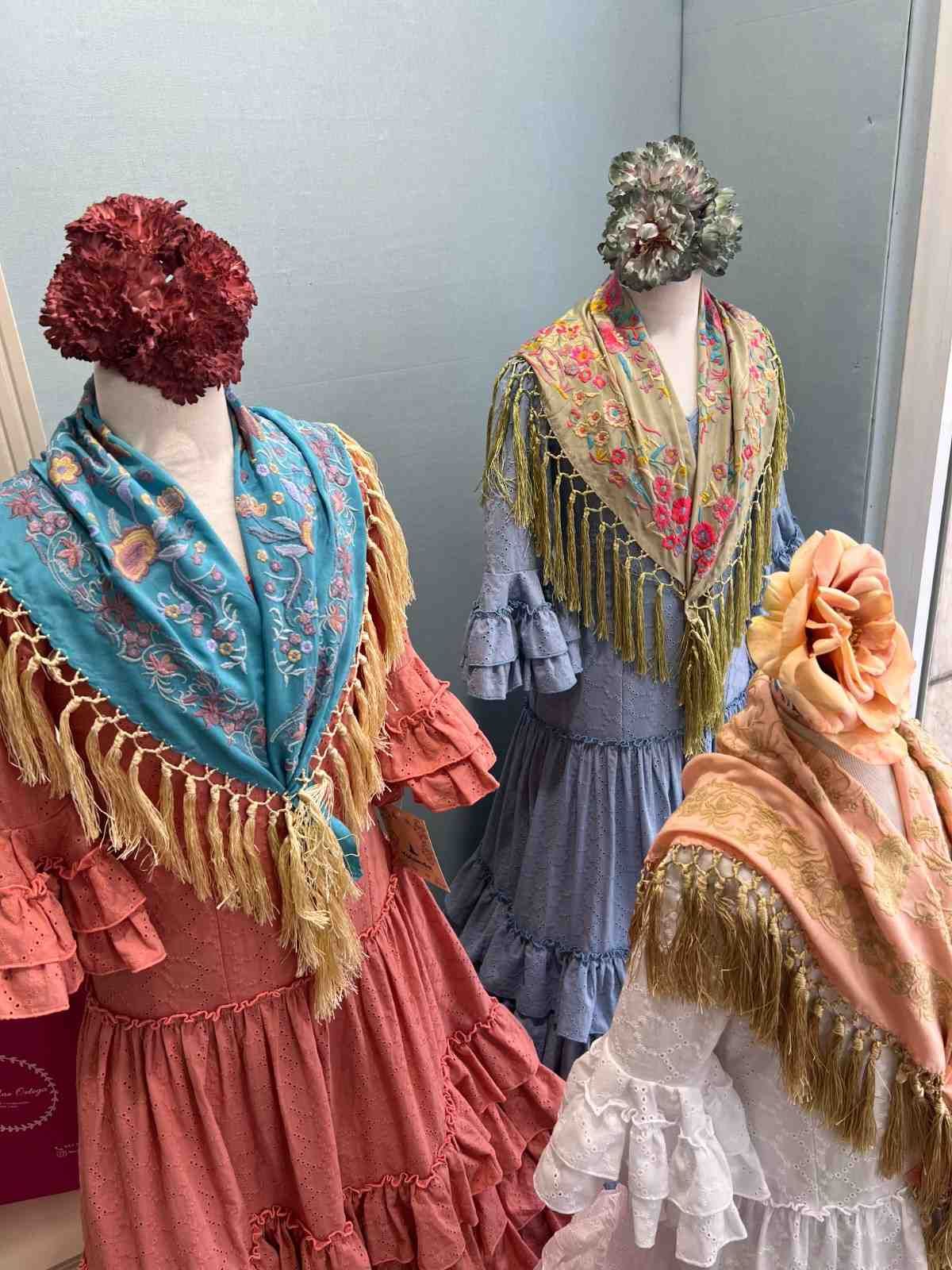 Tres maniquíes de estilo vintage exhiben chales coloridos y vestidos superpuestos en el escaparate de una tienda.