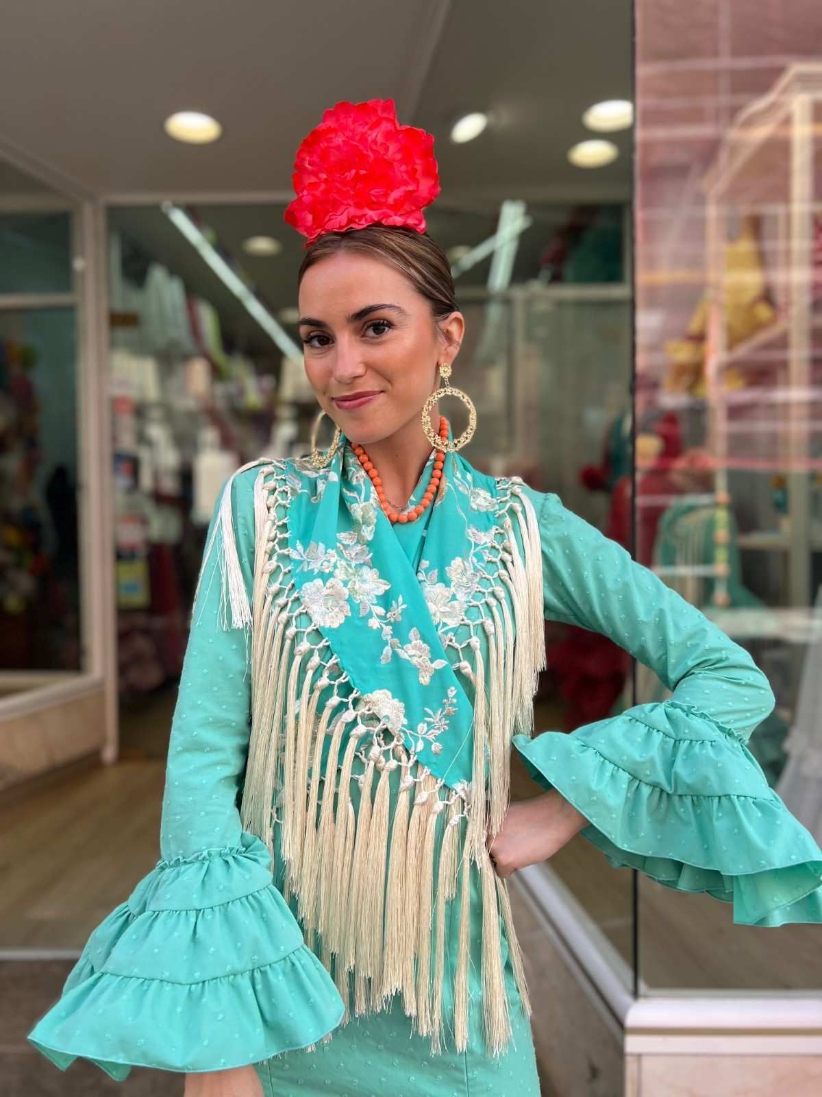 Mujer con vestido flamenco turquesa y tocado rojo de pie en la entrada de una tienda.