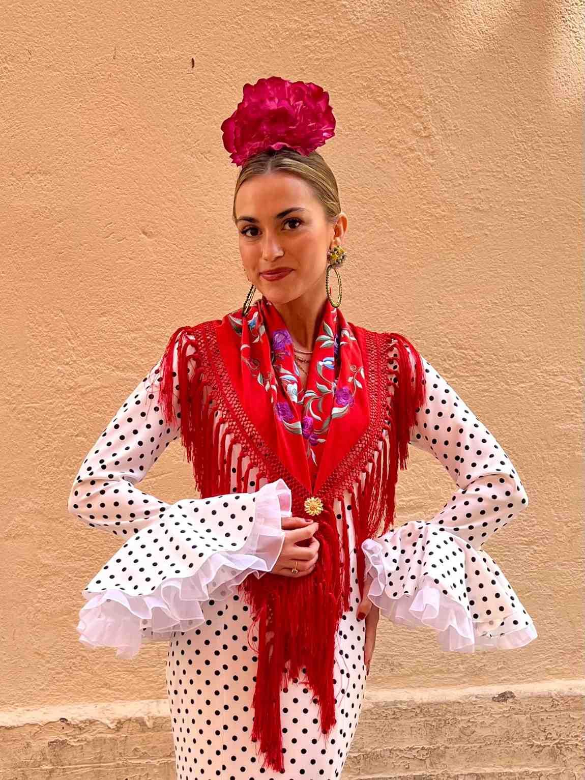 Mujer con vestido rojo con flecos, mangas blancas de lunares y tocado floral rosa, de pie contra una pared color melocotón.