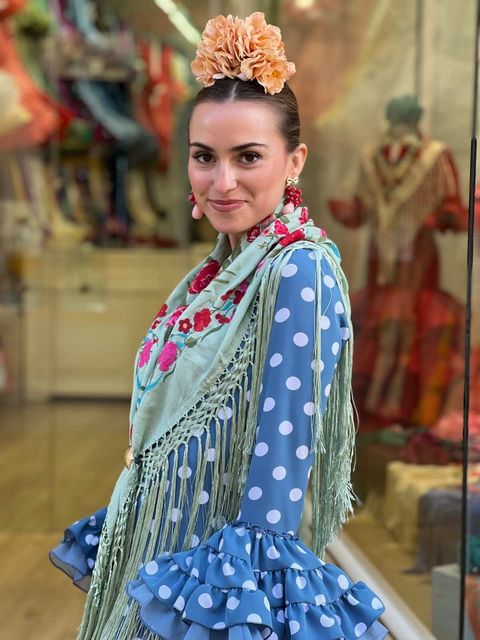 Mujer con vestido azul de lunares y bufanda floral de pie en el escaparate de una tienda.