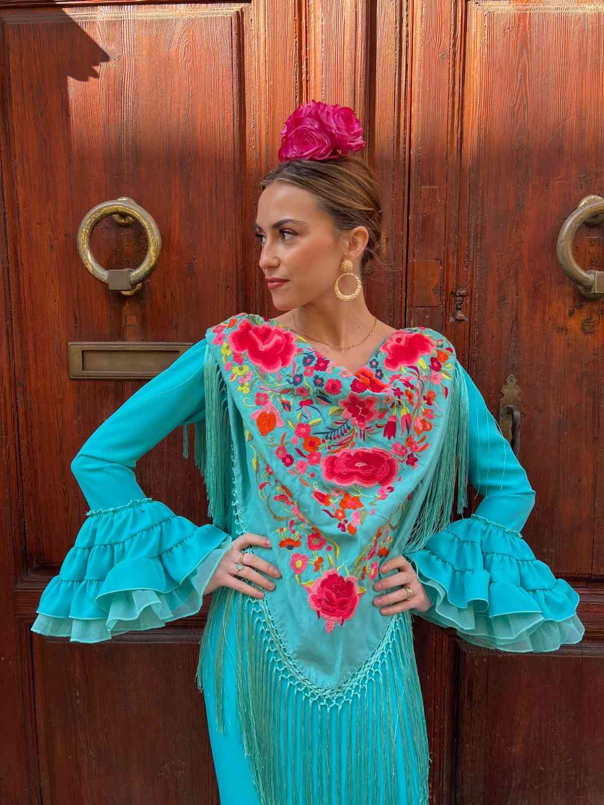 Mujer con vestido turquesa bordado y mangas con volantes, de pie frente a una puerta de madera tallada.