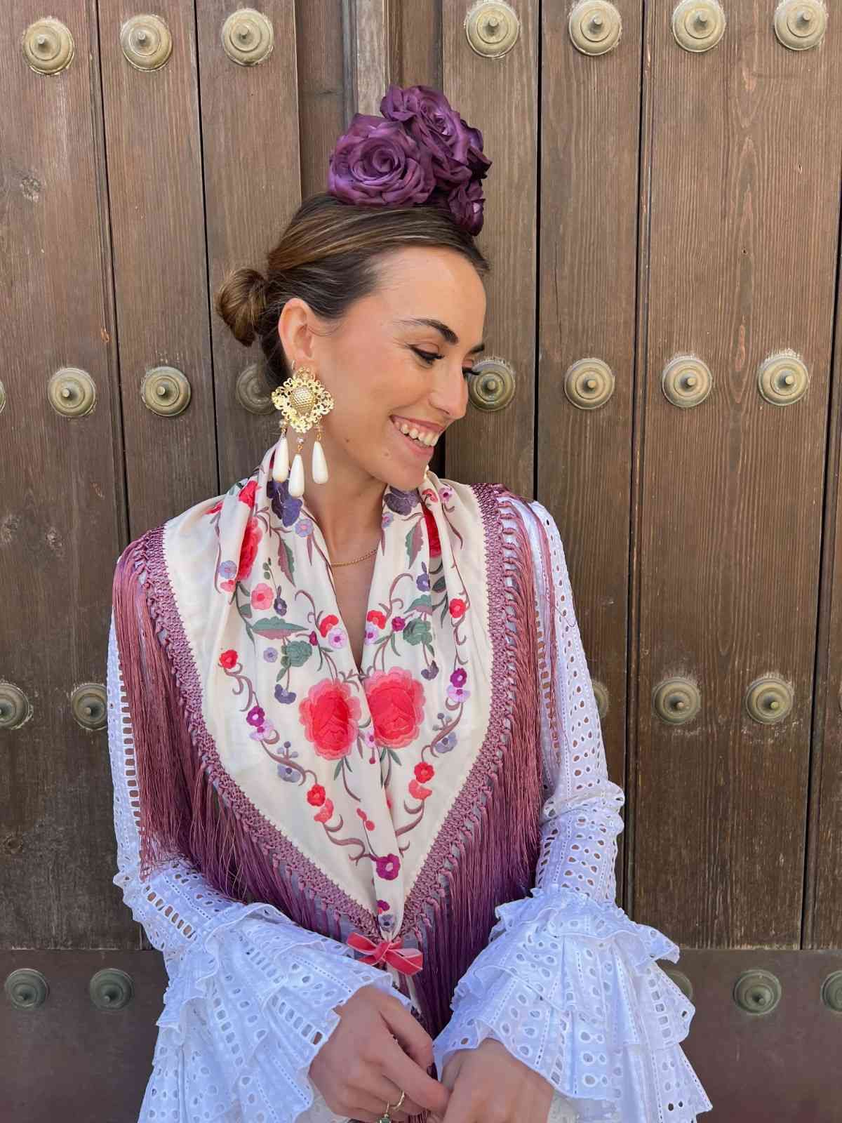 Mujer con blusa blanca y chaleco morado sonriendo frente a una puerta de madera con tachuelas metálicas decorativas.