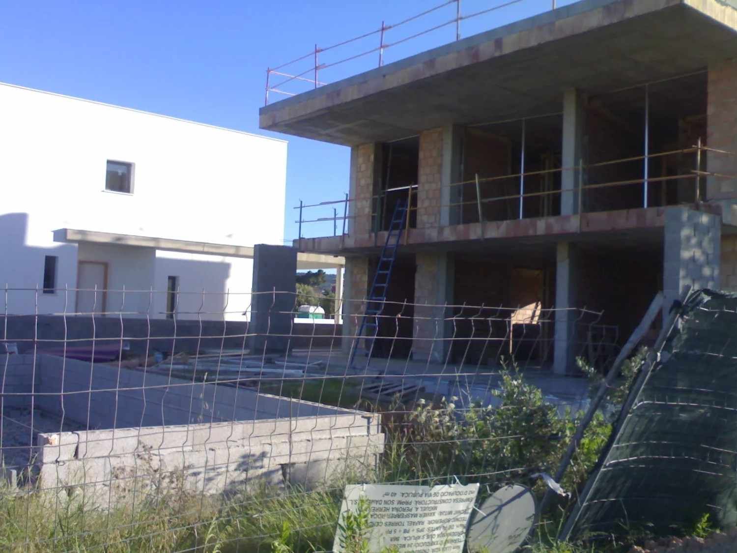 Edificio de hormigón en construcción detrás de un patio vallado, bajo un cielo azul despejado