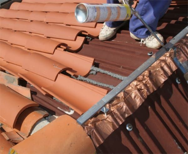 Obrero de techado reemplazando tejas de terracota en un tejado con un martillo y manos enguantadas.