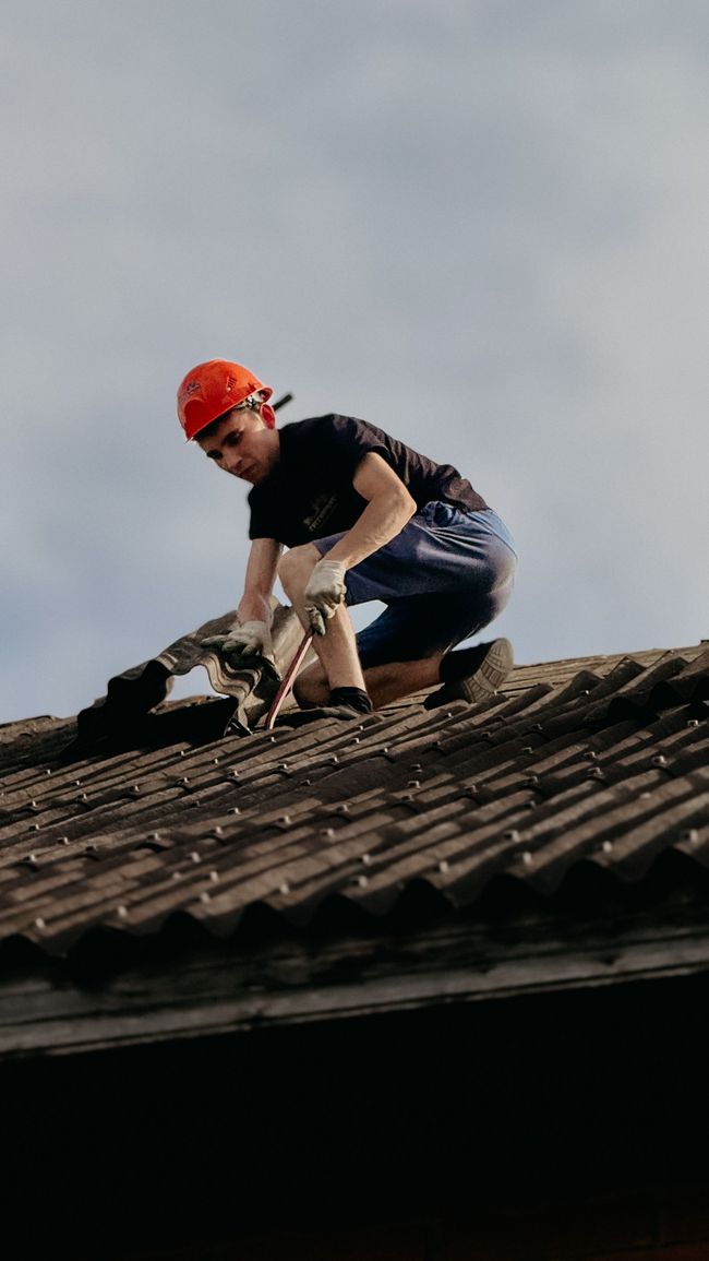 Un trabajador con casco rojo, agachado sobre un tejado de tejas, lo repara con herramientas.