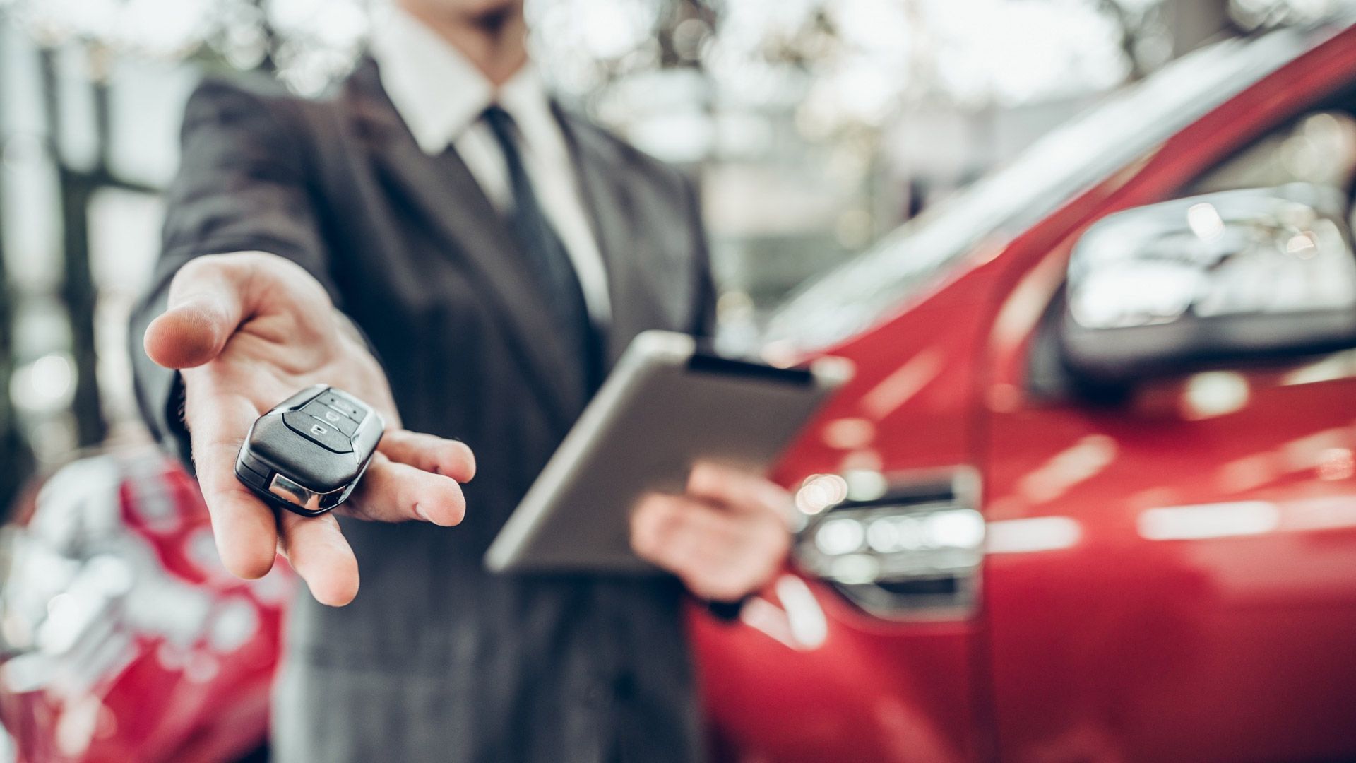Un hombre con traje sostiene una llave de coche delante de un coche rojo.