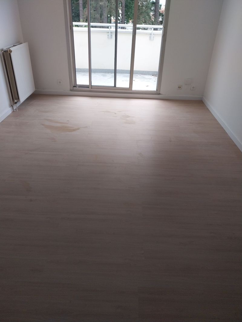 Pièce vide avec parquet, grande fenêtre et radiateur mural.